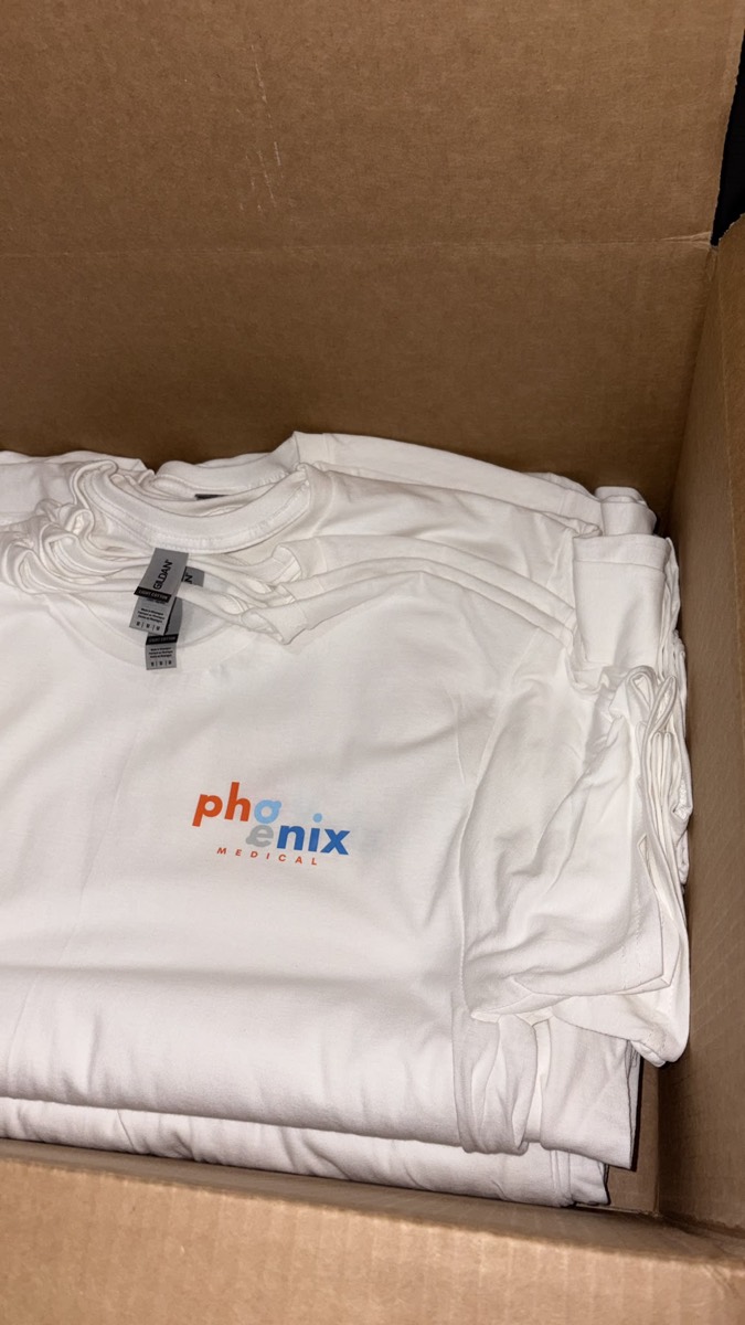Box of folded white tees with a small Phoenix Medical chest logo